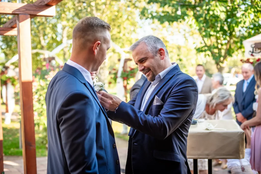 A ceremóniamester segít a vőlegénynek a kitűző felhelyezésében az esküvő előtti percekben. Bagi Péter – ceremóniamester