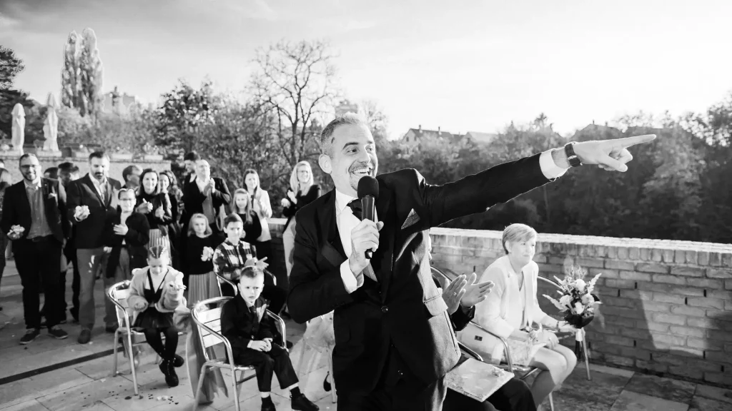A ceremóniamester az esküvői vendégek előtt áll, mikrofonnal a kezében, lendülettel irányítva az eseményeket. Bagi Péter – ceremóniamester